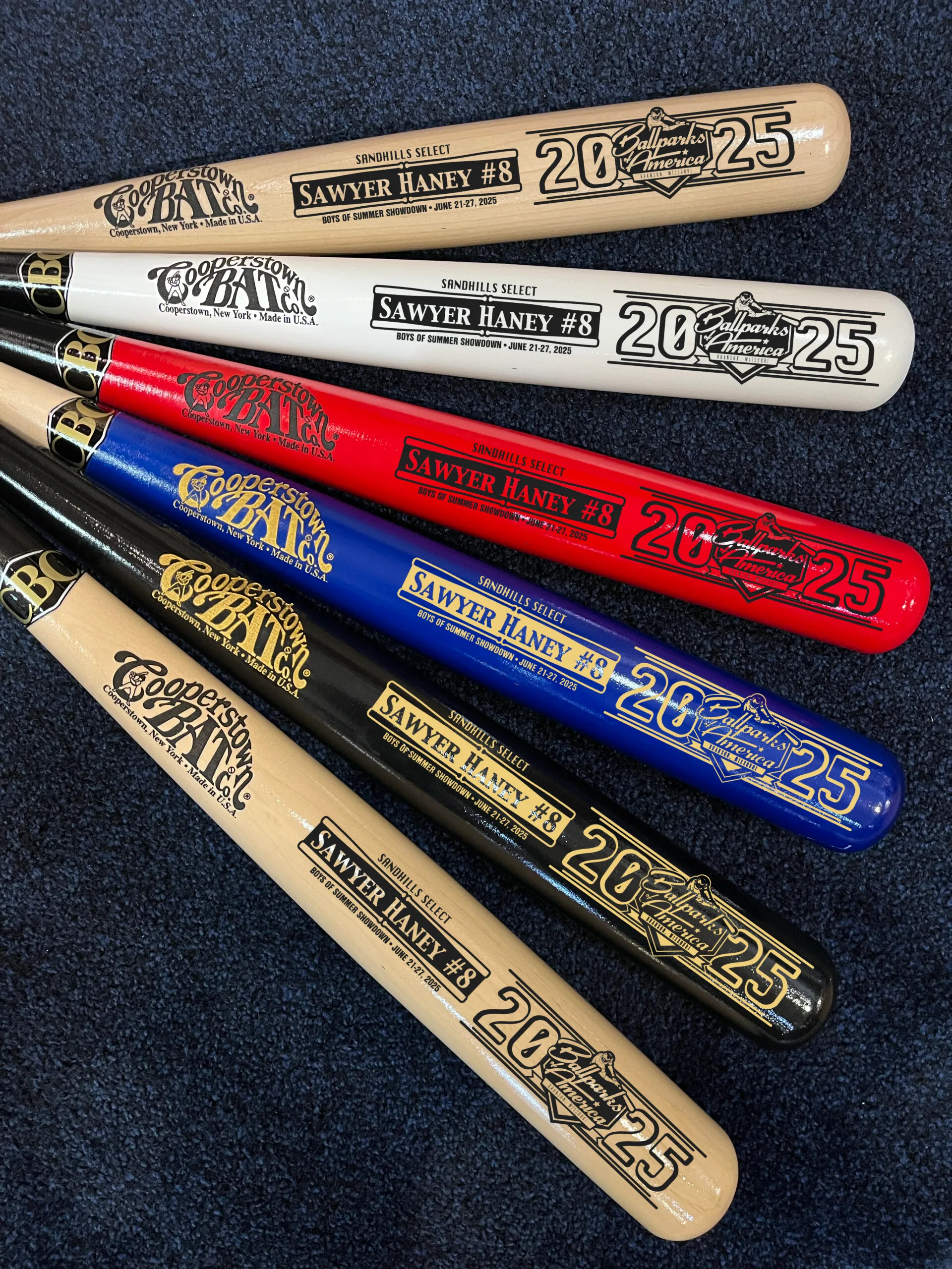 Colorful Cooperstown youth baseball bats on dark carpet.